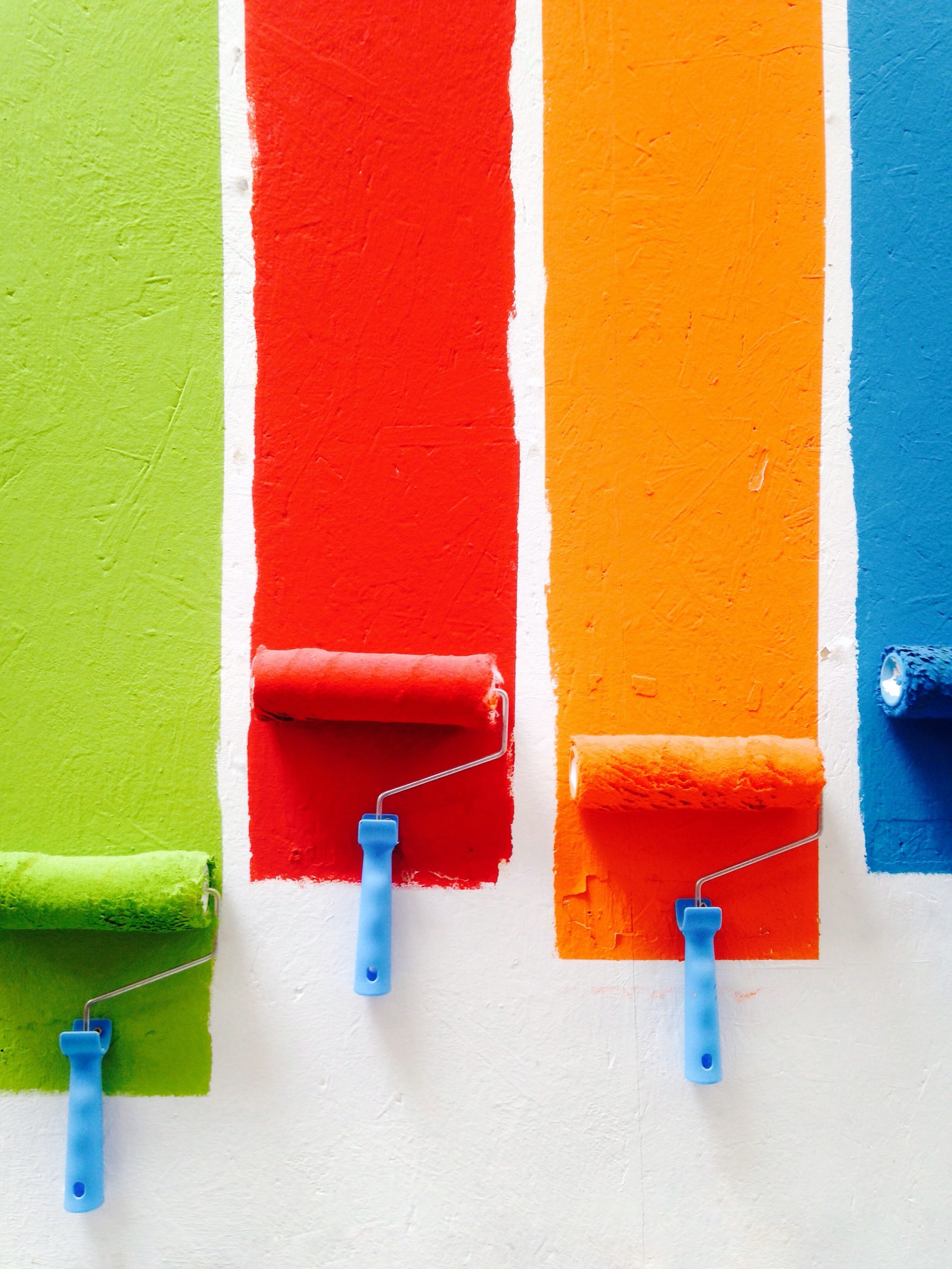 Four paint rollers painting on a white wall - green, red, orange, blue