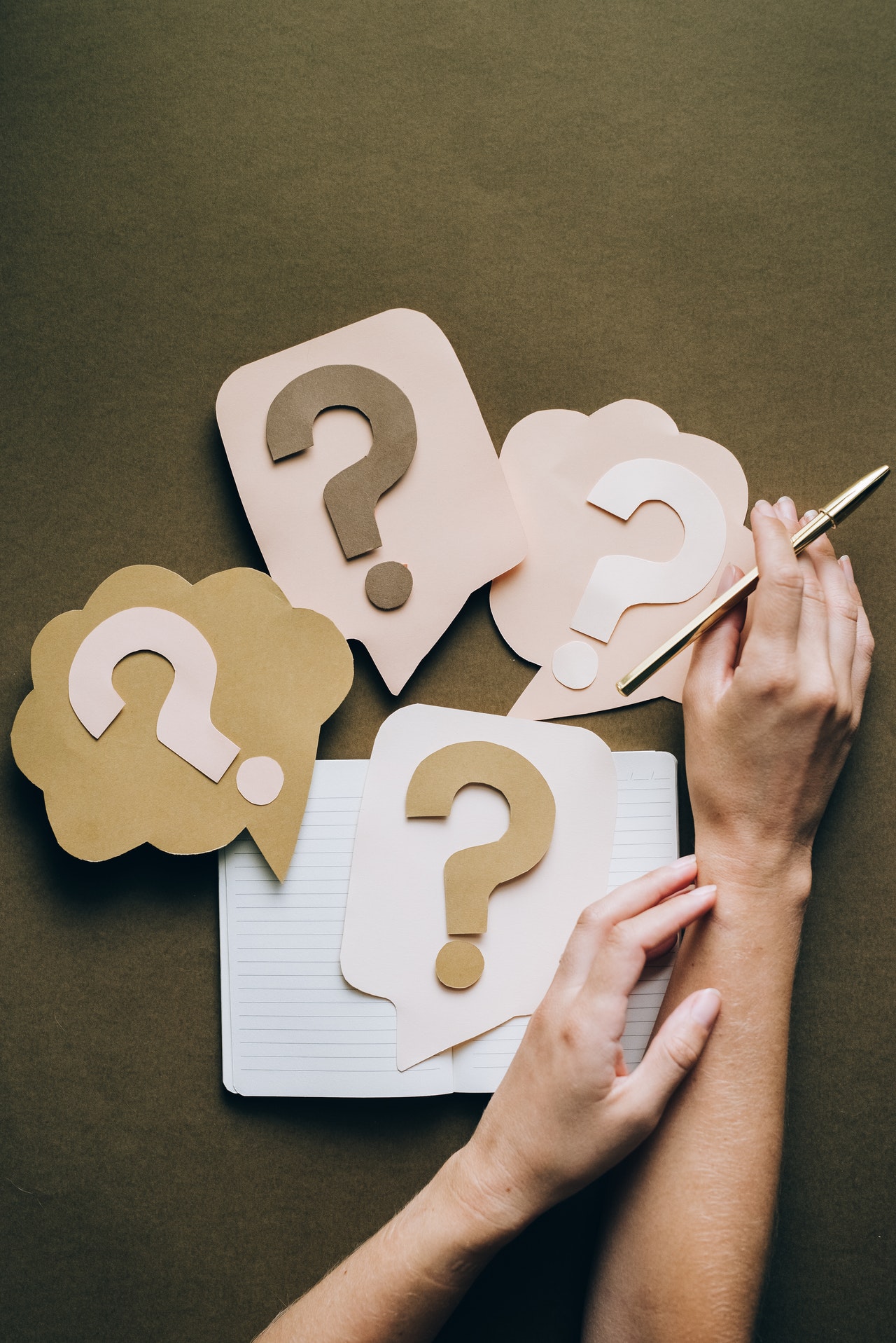 A top down view of a notebook, a pair of hands holding a pen and four cardboard cutouts of question marks