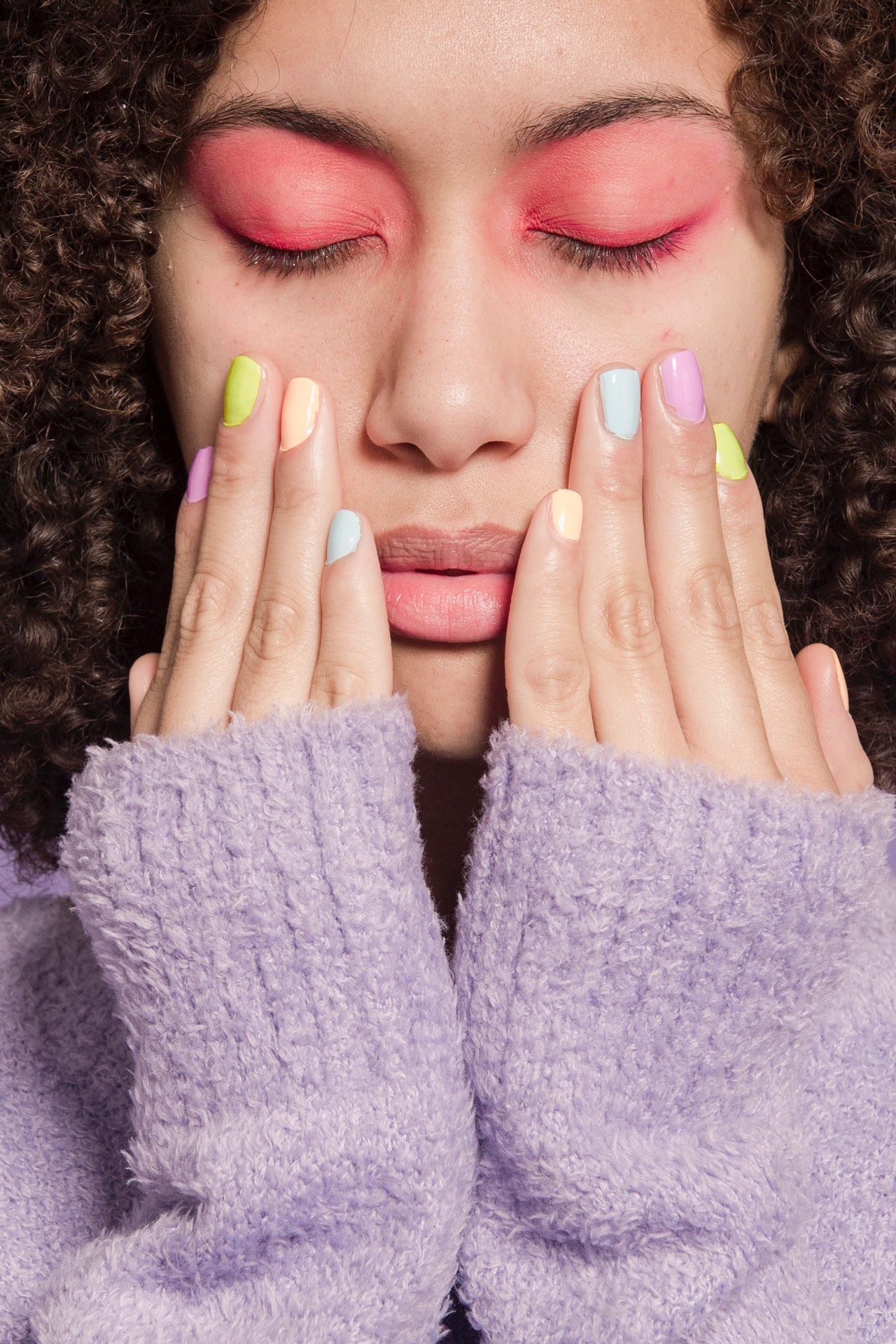 A woman with brown curly hair, closed eyes and pink eyeshadow. She's holding her hands up to her face and has different coloured pastel nail polish. She's wearing a lilac coloured woollen jumper.
