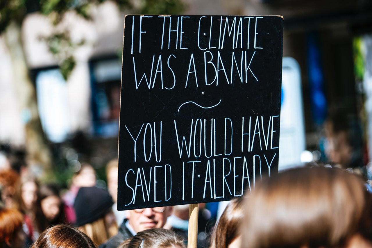 A black sign being held up in a crowd with white writing "If the climate was a bank you would have saved it already"